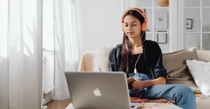 Jovem mulher sentada no sofá com fones de ouvido em frente ao seu laptop aprendendo como criar vídeo com roteiro com IA gratuito