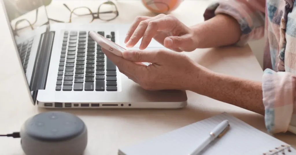 Pessoa usando IA generativa de imagens realistas no celular em frente ao seu laptop com óculos, caderno e caneta na mesa de trabalho