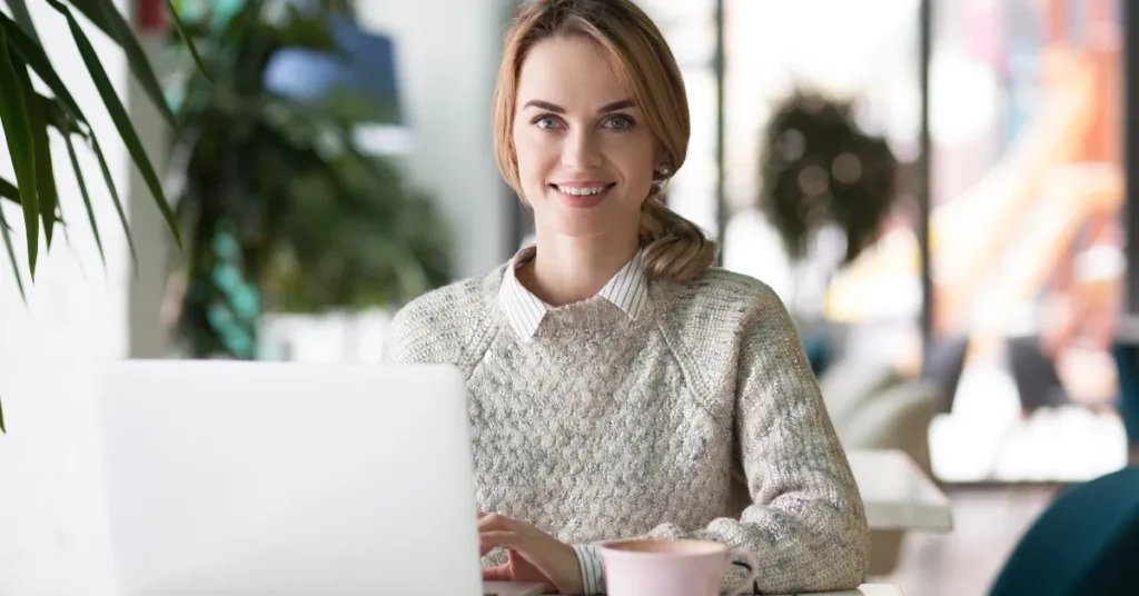 Mulher sentada em uma mesa em frente ao seu laptop olhando de frente e sorrindo porque aprendeu a usar IA para criar slides gratuitos