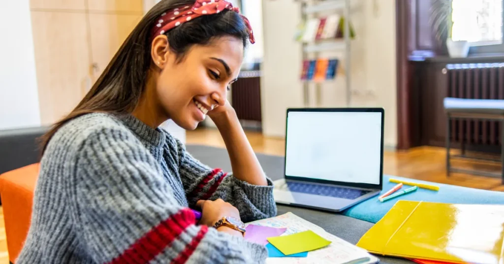 Jovem mulher sentada à mesa usando IA para estudos do seu dia a dia com laptop, caderno, pastas e post-its