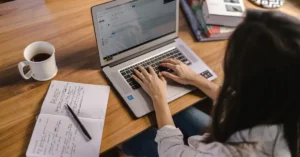 Mulher sentada em mesa de madeira com laptop, café, caderno e caneta usando IA para estudar