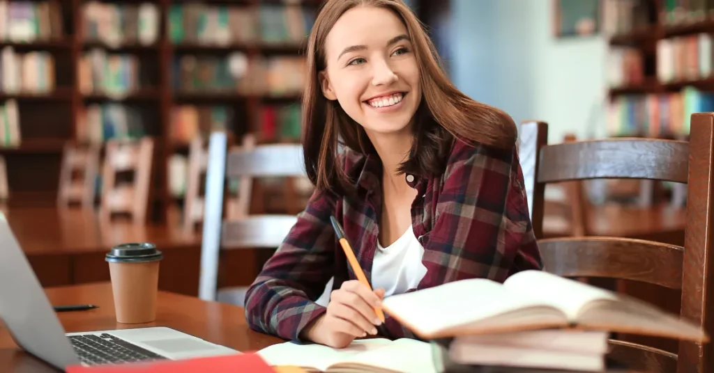 Jovem mulher sentada em uma biblioteca com laptop, livros e café sorrindo porque aprendeu os benefícios da IA para os estudantes